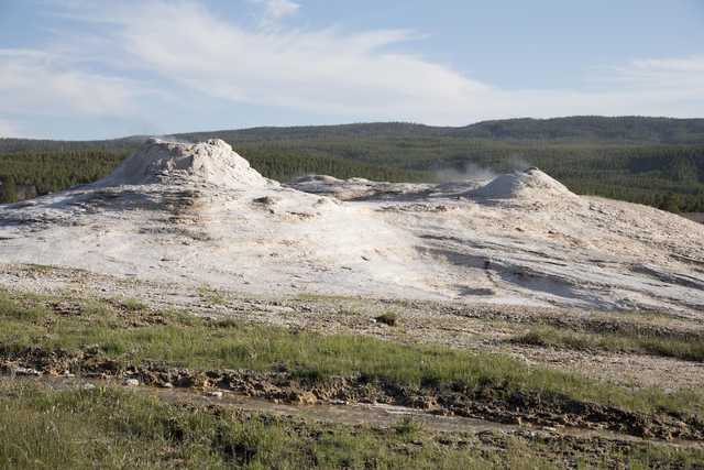A cluster of geysers and hot springs is visible in a grassy area with trees in the background.