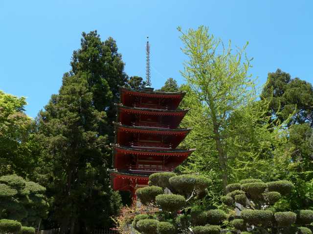 A traditional Japanese temple with multiple tiers and a red roof nestled among lush greenery including tall trees and manicured shrubs.