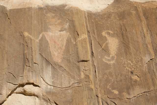 A rocky cliff face features two distinct petroglyphs, one resembling an abstract figure and the other a stylized animal with curved horns.