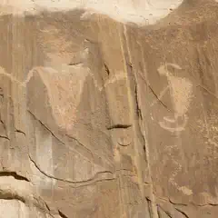 A rocky cliff face features two distinct petroglyphs, one resembling an abstract figure and the other a stylized animal with curved horns.