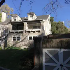 An off-white house with gray shingles and white trim with a chimney, a covered porch, and a staircase leading to a second floor balcony.