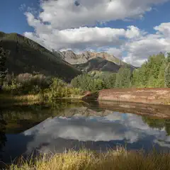 A pond reflects a forested mountain range under a blue sky with scattered clouds.