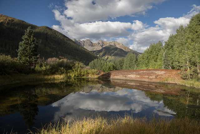 A pond reflects a forested mountain range under a blue sky with scattered clouds.