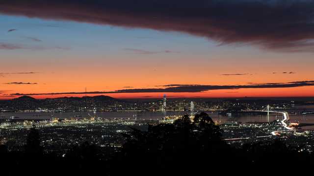 A panoramic view of a coastal city at dusk, featuring a prominent bridge spanning a bay, illuminated urban areas, and a distant mountainous horizon.