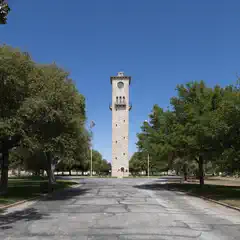 A tall stone clock tower stands at the end of a tree-lined street, with a clear blue sky above.