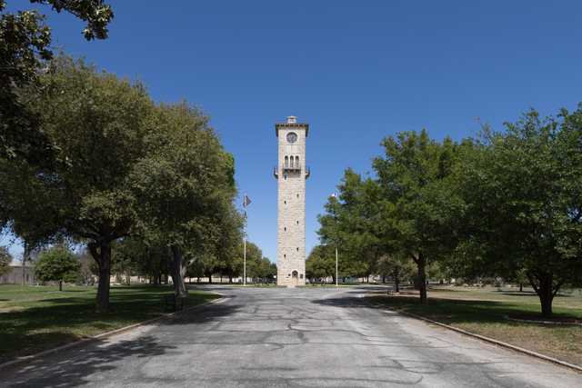A tall stone clock tower stands at the end of a tree-lined street, with a clear blue sky above.