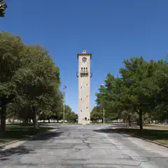 A tall stone clock tower stands at the end of a tree-lined street, with a clear blue sky above.