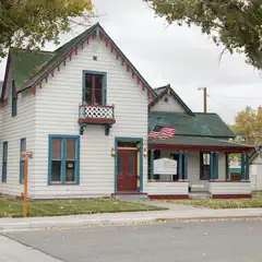 A white house with green trim and a red door, featuring a flag on the roof and a sign in front of it, sitting next to trees and a street with a sidewalk.