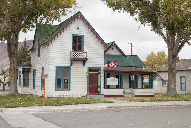 A white house with green trim and a red door, featuring a flag on the roof and a sign in front of it, sitting next to trees and a street with a sidewalk.