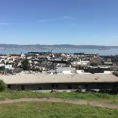 A low, dark building sits in the foreground with a dense residential area extending toward a body of water and distant hills.