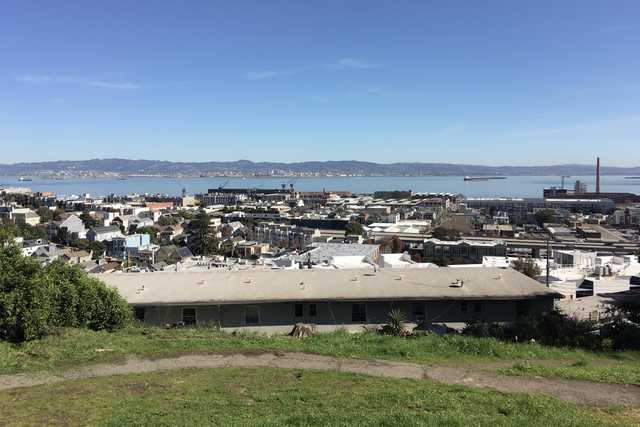 A low, dark building sits in the foreground with a dense residential area extending toward a body of water and distant hills.