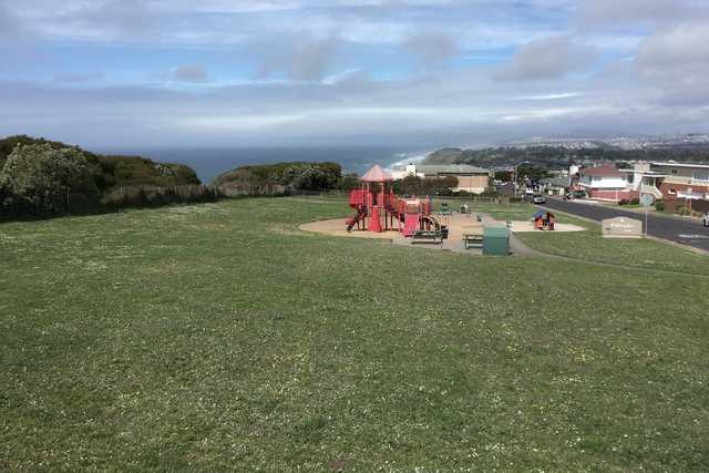 A grassy field slopes down to a playground and a residential neighborhood overlooking the ocean.