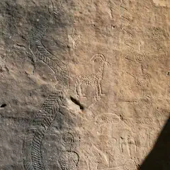 A group of petroglyphs on a flat rock face includes geometric designs, human and animal figures, and a large, detailed snake.