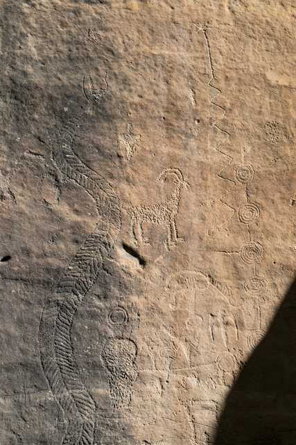 A group of petroglyphs on a flat rock face includes geometric designs, human and animal figures, and a large, detailed snake.