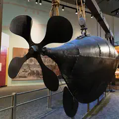 A small, black submarine with a large, four-bladed propeller on display in a museum.