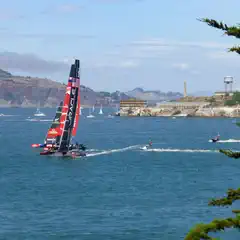 A racing sailboat with a black hull and red sail is on the water. Smaller sailboats and jet skis are visible in the distance near a rocky coastline and hills.