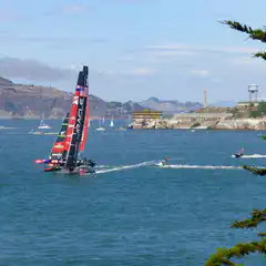 A racing sailboat with a black hull and red sail is on the water. Smaller sailboats and jet skis are visible in the distance near a rocky coastline and hills.