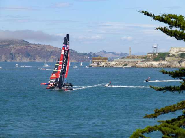 A racing sailboat with a black hull and red sail is on the water. Smaller sailboats and jet skis are visible in the distance near a rocky coastline and hills.