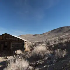 An abandoned wooden shack sits on a rocky hillside, surrounded by sparse vegetation under a clear sky.