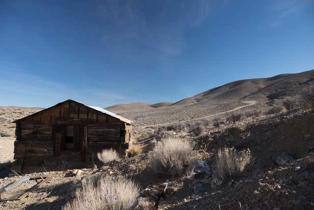 An abandoned wooden shack sits on a rocky hillside, surrounded by sparse vegetation under a clear sky.