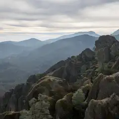 A rocky outcrop with sparse vegetation overlooks a range of misty mountains under a cloudy sky.