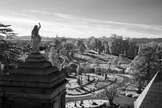 A statue stands on a stone base overlooking a cemetery with a city skyline in the distance.