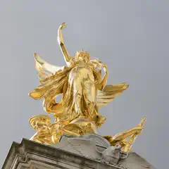 A golden statue of a winged figure with outstretched arms stands atop a stone monument against a pale sky.