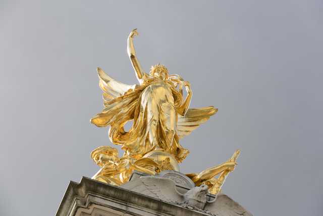 A golden statue of a winged figure with outstretched arms stands atop a stone monument against a pale sky.
