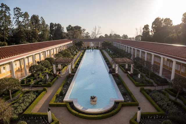 A long rectangular pool is surrounded by a garden with trimmed hedges and trees, flanked by two rows of white columns supporting red-tiled roofs.