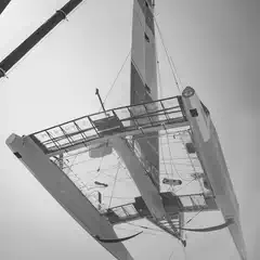 A modern racing sailboat is suspended in the air, showcasing its sleek, high-tech design and complex rigging.