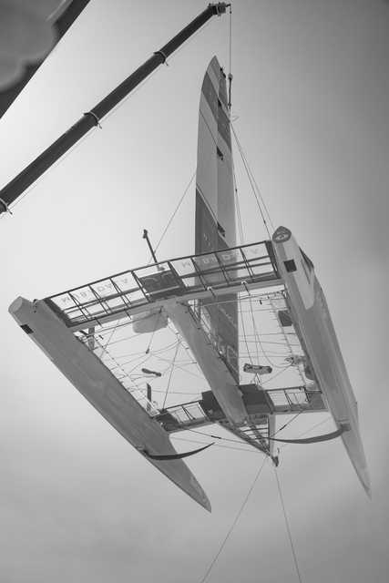 A modern racing sailboat is suspended in the air, showcasing its sleek, high-tech design and complex rigging.