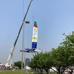 A large crane is lifting a colorful sailboat into the air, with trees and buildings visible in the background under a clear sky.