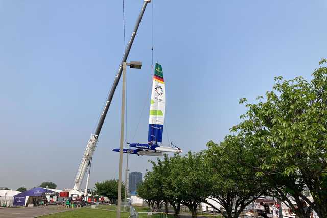 A large crane is lifting a colorful sailboat into the air, with trees and buildings visible in the background under a clear sky.