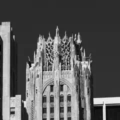 A tall, ornate building with Gothic-inspired spires and a central sculpted figure rises above surrounding structures against a dark sky.