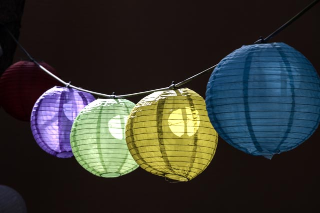 A string of paper lanterns in various colors hangs from a wire against a dark brown background.