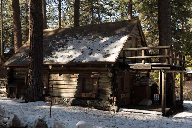 A cabin with a brown wooden exterior and stone foundation is surrounded by tall trees.
