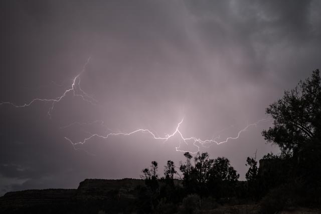 A dramatic lightning bolt illuminates a dark, stormy sky over a silhouette of trees and a rocky landscape.