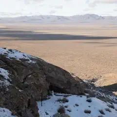 A rocky hillside with sparse vegetation and patches of snow overlooking a vast desert landscape.