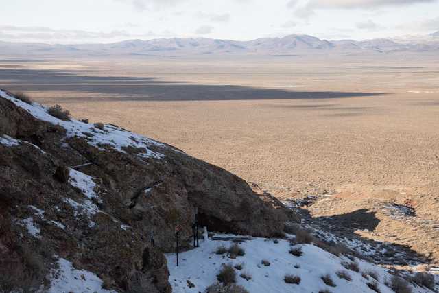 A rocky hillside with sparse vegetation and patches of snow overlooking a vast desert landscape.