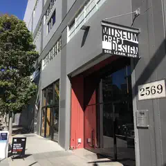 Gray building with large windows, red door, sign that says "Museum of Art & Design" and number 2569 on the side, partly shaded by green trees.