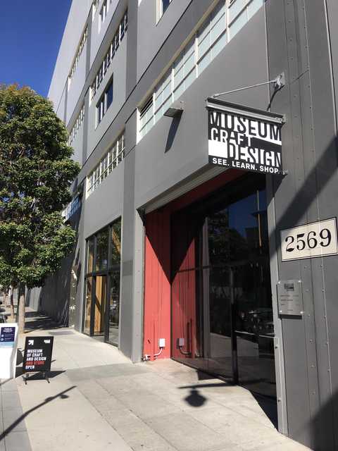 Gray building with large windows, red door, sign that says "Museum of Art & Design" and number 2569 on the side, partly shaded by green trees.