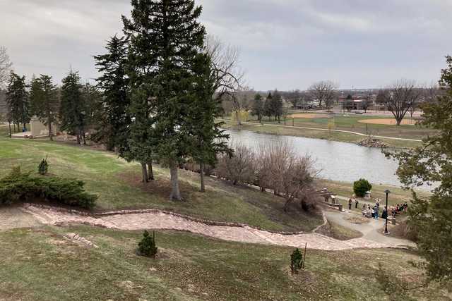 A path leads down to a lake, lined with trees and grassy areas, where several people are gathered near water's edge.