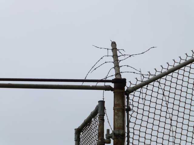 A rusted metal fence topped with barbed wire, standing against a gray sky.