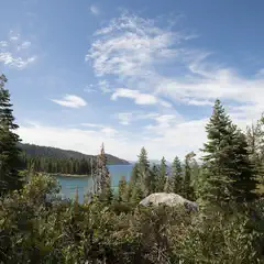 A serene lake surrounded by dense evergreen trees under a bright blue sky with scattered clouds.