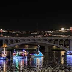 A bridge at night, decorated with lights and illuminated by moonlight, over water where boats are adorned with colorful lights.