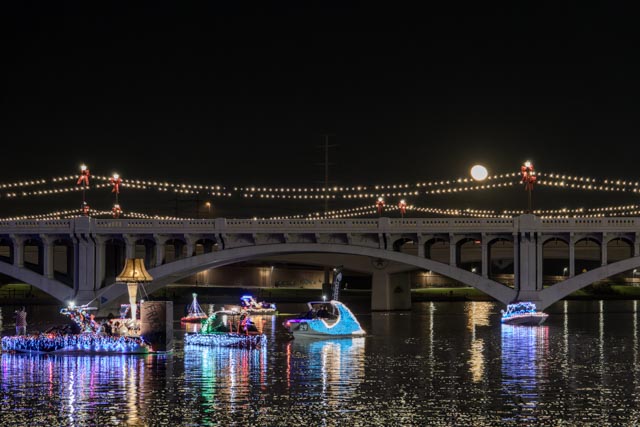 A bridge at night, decorated with lights and illuminated by moonlight, over water where boats are adorned with colorful lights.