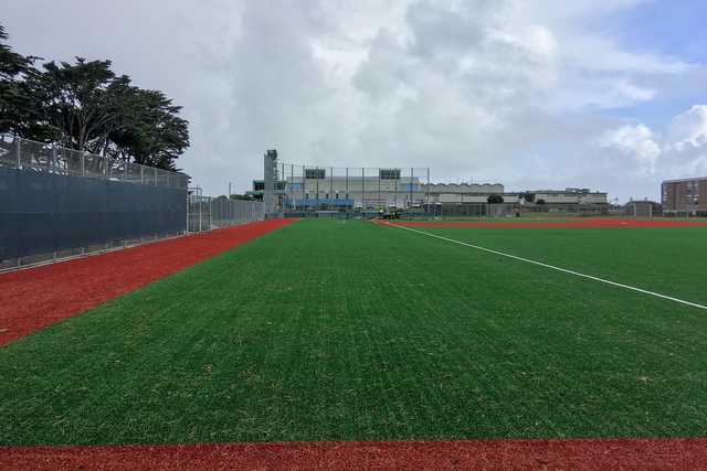 A green baseball field has a red base path and a dark fence running along the left side, with buildings visible in the distance under a cloudy sky.