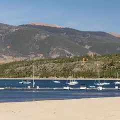 A mountainous coastline is visible, with several boats docked at the pier. The water is calm and a sandy beach can be seen on the right side of the image.