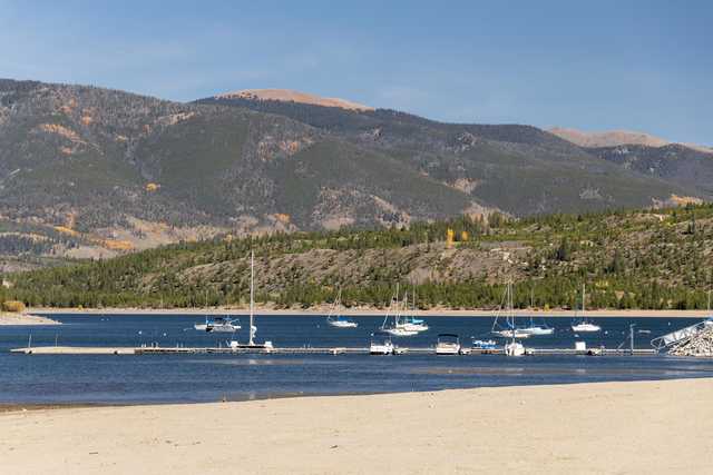 A mountainous coastline is visible, with several boats docked at the pier. The water is calm and a sandy beach can be seen on the right side of the image.