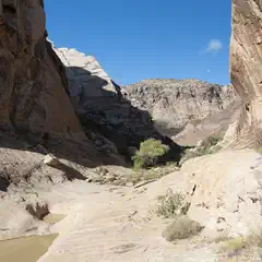 A rocky canyon with a narrow stream flowing through it, surrounded by rugged terrain under a clear blue sky.